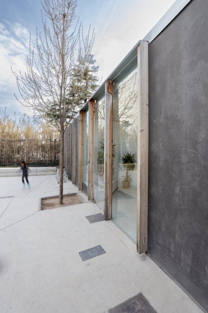 Fachada acristalada de la ludoteca con montantes de madera y un niño jugando en la plaza